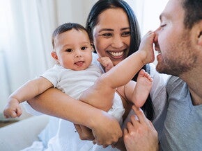 Mamá carga bebé sonriendo. Bebé tiene el pie en la nariz de papá. Mamá carga bebé sonriendo. Bebé tiene el pie en la nariz de papá.