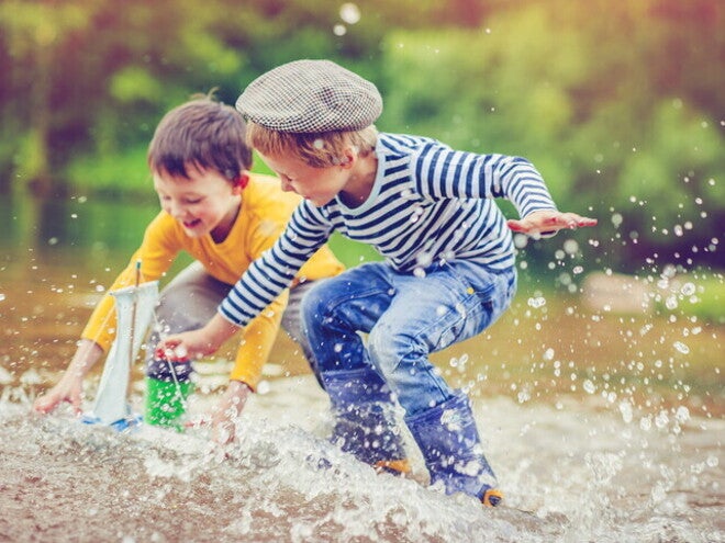 Dos niños pequeños juegan juntos en el agua, lanzando y persiguiendo un barco de juguete, mostrando interacción social en un entorno natural.