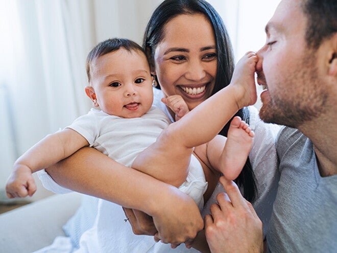 Mamá carga bebé sonriendo. Bebé tiene el pie en la nariz de papá. Mamá carga bebé sonriendo. Bebé tiene el pie en la nariz de papá.