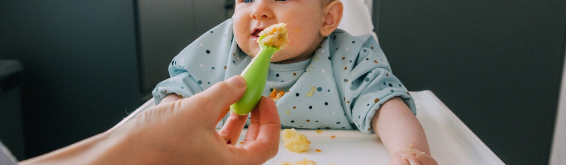 Mano de mamá dando de comer con una cuchara a su bebé que está sentado en silla alta. Alimentación complementaria del bebé.